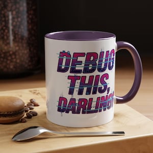 May include: White ceramic mug with a purple interior and handle. The mug features the text "DEBUG THIS, DARLING" in a purple and blue gradient font. A spoon and a macaron are on a wooden surface.