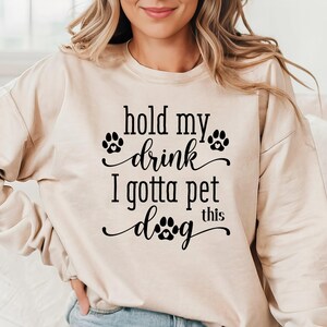 Può includere: Felpa beige con la scritta nera "hold my drink I gotta pet this dog". Il design include impronte di zampe. La felpa è di colore neutro e ha maniche lunghe.