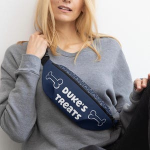 Può includere: Un marsupio blu navy con la scritta "Duke's Treats" in bianco, insieme a grafiche di ossa bianche. La borsa ha un cinturino nero e un motivo lungo la cerniera.
