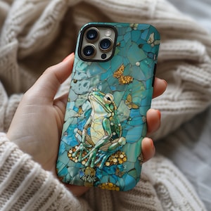 May include: A blue and green phone case with a mosaic design featuring a frog sitting on a mushroom and butterflies. The case has a cutout for the camera lens.