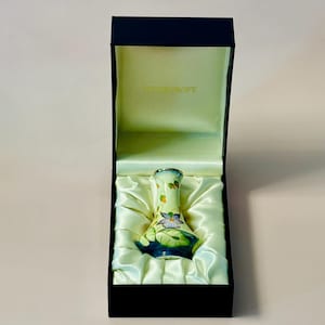 May include: A small, hand-painted porcelain vase with floral designs in shades of blue, green, and yellow. The vase is displayed in a black velvet box with a white satin lining.