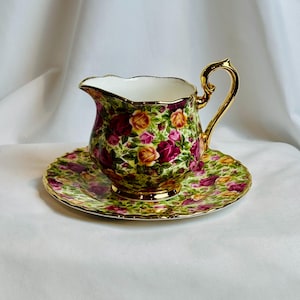 May include: A porcelain creamer pitcher and matching saucer set featuring a vibrant floral pattern of red, pink, and yellow roses on a green leafy background. The creamer has a scalloped rim, a gold-trimmed handle, and a gold-finished base.