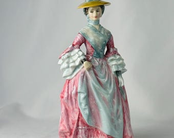 以下が含まれることがあります： 歴史的なドレスを着た女性の磁器製フィギュア。フィギュアは黄色の帽子、フリル袖の青とピンクのドレス、黒い靴を着用しています。フィギュアは複雑なディテールで塗装されています。