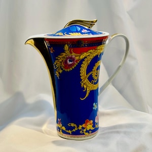 May include: A tall porcelain coffee pot featuring a vibrant royal blue body adorned with intricate gold filigree, red accents, and colorful floral motifs. The lid is topped with a gold finial, and the spout and handle are finished in gold.