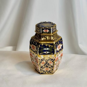 May include: A hexagonal porcelain ginger jar featuring a deep blue base color with intricate gold filigree patterns and floral motifs in shades of orange and white. The jar includes a matching removable lid with a gold-trimmed hexagonal top.
