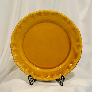 May include: A round, mustard yellow ceramic pie dish with a smooth, glazed finish and a decorative, scalloped, ruffled edge. The dish is displayed upright on a small, black, floral-patterned stand, showcasing its vibrant color and texture.