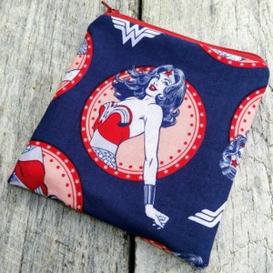 May include: A blue zippered pouch with a Wonder Woman print. The print features Wonder Woman in a red and white costume with a star-studded background. The pouch has a red zipper and a white Wonder Woman logo.