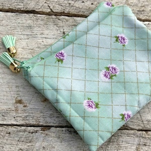 May include: A light green zippered pouch with a gold zipper and tassels. The pouch has a white and gold grid pattern with small pink flowers.