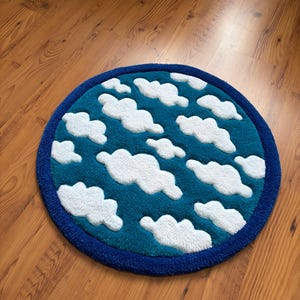 May include: A round rug with a blue border and a teal center. The teal area features white, fluffy cloud shapes. The rug is designed to resemble a sky with clouds.