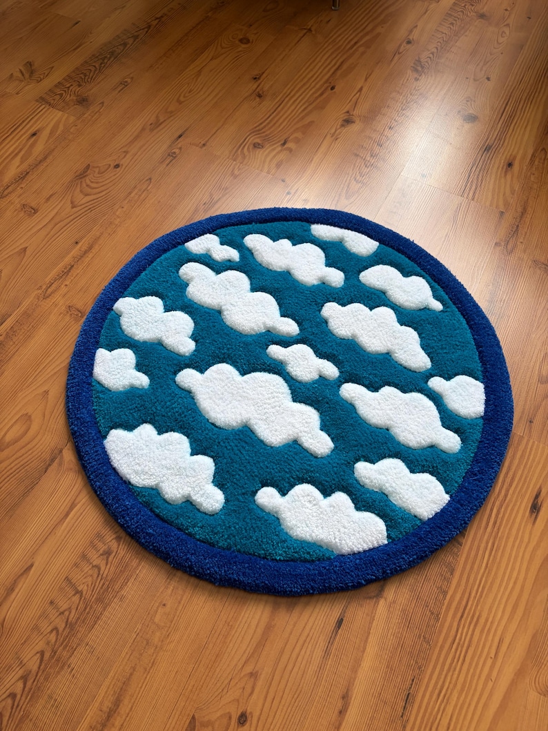 May include: A round rug with a blue border and a teal center. The teal area features white, fluffy cloud shapes. The rug is designed to resemble a sky with clouds.