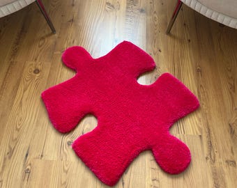 May include: A bright red fluffy rug shaped like a jigsaw piece. The rug is on a wooden floor.
