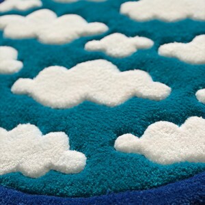 May include: A round rug with a sky and cloud design. The rug features a teal blue background with white, fluffy cloud shapes. A dark blue border frames the design.