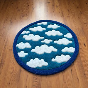 May include: A round rug with a sky and cloud design. The rug features a teal blue center with white fluffy cloud shapes and a dark blue border. The rug is placed on a wooden floor.