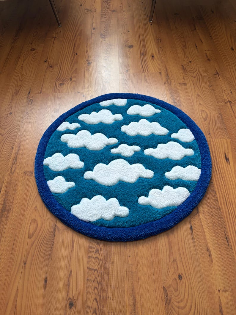 May include: A round rug with a sky and cloud design. The rug features a teal blue center with white fluffy cloud shapes and a dark blue border. The rug is placed on a wooden floor.