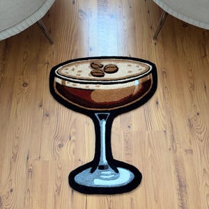 May include: A rug shaped like a cocktail glass, featuring a brown drink with a tan foam and three coffee beans. The glass has a black outline and a light blue stem and base. The rug is on a wooden floor.