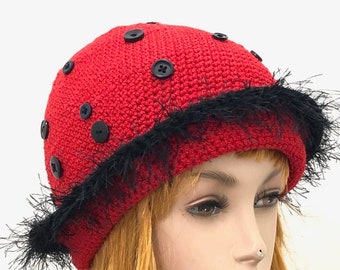 May include: A red crocheted hat with black buttons and a black fringe trim. The hat is on a mannequin head with long blonde hair. The hat has a rounded crown and a small brim.