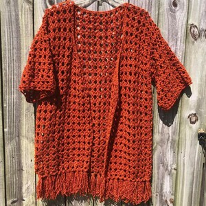 May include: An open-weave, rust-colored crochet cardigan with short sleeves and a fringed hem. The handmade garment hangs on a clear plastic hanger against a weathered wooden fence. The cardigan's texture and color are prominent.