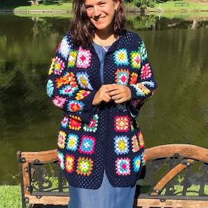 May include: A woman wearing a blue crochet cardigan with colorful square patterns. The cardigan is open and the woman is standing in front of a wooden bench near a pond.