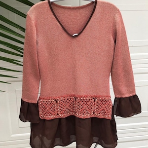 May include: A long-sleeved, pink sweater with a brown crocheted trim and a brown sheer overlay. The sweater has a V-neck and ruffled sleeves.
