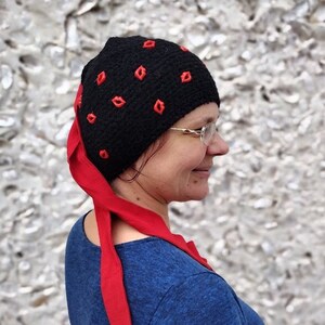 May include: A black knitted hat with red lips embroidered on it. The hat has a long red tie that hangs down the back of the neck.