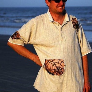 May include: A man wearing a light beige polo shirt with a spiderweb design on the front. The shirt has several rips and tears. The man is standing on a beach with the ocean in the background.