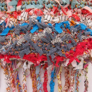 May include: A colorful woven rug with a fringe made of various fabrics, including denim, red, blue, and white. The rug is made with a variety of textures and patterns.