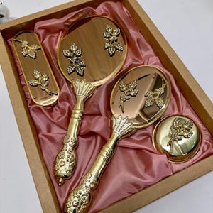 May include: A gold-toned vanity set with a hand mirror, a small round mirror, and a rectangular tray, all adorned with floral and crystal accents. The set is presented in a brown box with a pink satin lining, creating an elegant presentation.