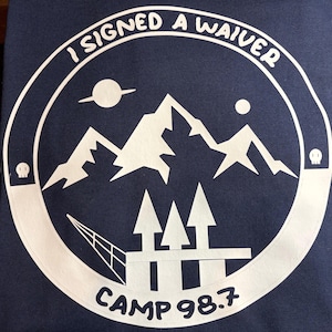 May include: Navy blue t-shirt with a white graphic. The graphic features a mountain range, trees, a planet with a ring, and the text "I SIGNED A WAIVER" and "CAMP 98.7" in a circular design. The design includes two skull icons.