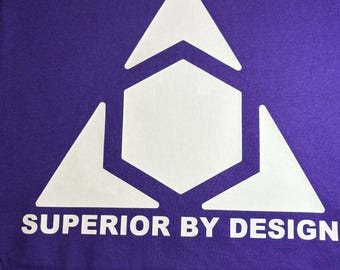 May include: A purple t-shirt featuring a white geometric design composed of triangles and a hexagon. The text "SUPERIOR BY DESIGN" is printed in white below the graphic. The shirt is made of a soft fabric.