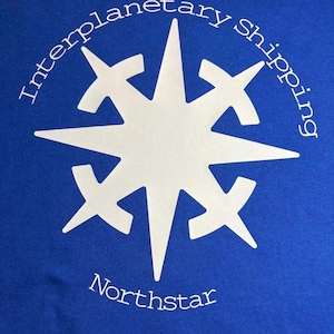 May include: Royal blue t-shirt with a white starburst design. The words "Interplanetary shipping" are arched above the starburst, and "Northstar" is below. The design is centered on the shirt.