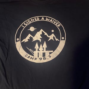 May include: Navy blue t-shirt with a white circular graphic. The graphic features mountains, arrows, and the text "I SIGNED A WAIVER" and "CAMP 98.7". The shirt is made of a soft material.