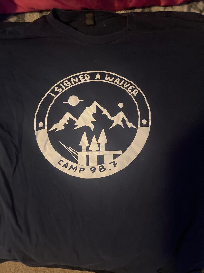 May include: Navy blue t-shirt with a white circular graphic. The graphic features mountains, arrows, and the text "I SIGNED A WAIVER" and "CAMP 98.7". The shirt is made of a soft material.