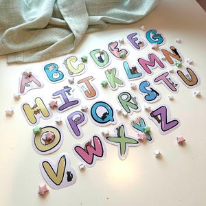 May include: A set of colorful alphabet stickers with cute cats on each letter. The letters are arranged in alphabetical order and are surrounded by small white stars.