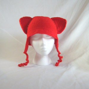 以下が含まれることがあります： 大きな耳の形をしたフラップと長いカールした紐が付いた、鮮やかな赤色の手編みの帽子。白いマネキンの頭に飾られており、手作りの質感とユニークなデザインが際立っています。この気まぐれな帽子は、コスチュームや普段使いに最適です。
