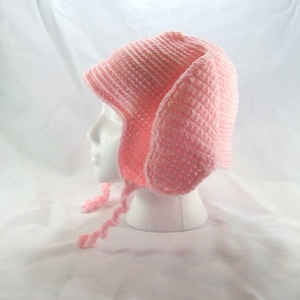 May include: A light pink, hand-crocheted hat with long ear flaps and ties. The hat is displayed on a white mannequin head against a white background. The knitted texture of the hat is clearly visible.