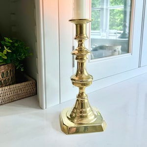 May include: A lit, ivory-coloured candle sits atop a gold-coloured candlestick. The candlestick has a square base and a tiered design. The flame is bright yellow. The candlestick is approximately 30 cm tall.