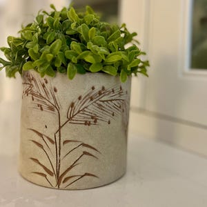 May include: A beige ceramic plant pot with a brown botanical design, filled with bright green artificial foliage. The pot is cylindrical and sits on a white surface. The design features stylized grass or wheat, adding a touch of nature.