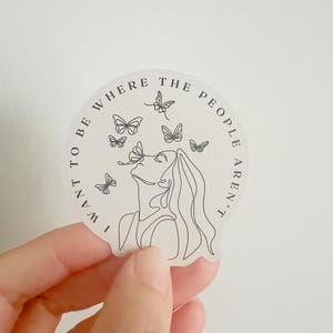 May include: A round sticker with a line drawing of a person with butterflies. The sticker has the text "I WANT TO BE WHERE THE PEOPLE AREN'T" in a circular pattern around the image.