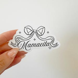 May include: A white sticker featuring a black bow and the word "Mamacita" in cursive. The design includes a ribbon and four star shapes. A decorative sticker.