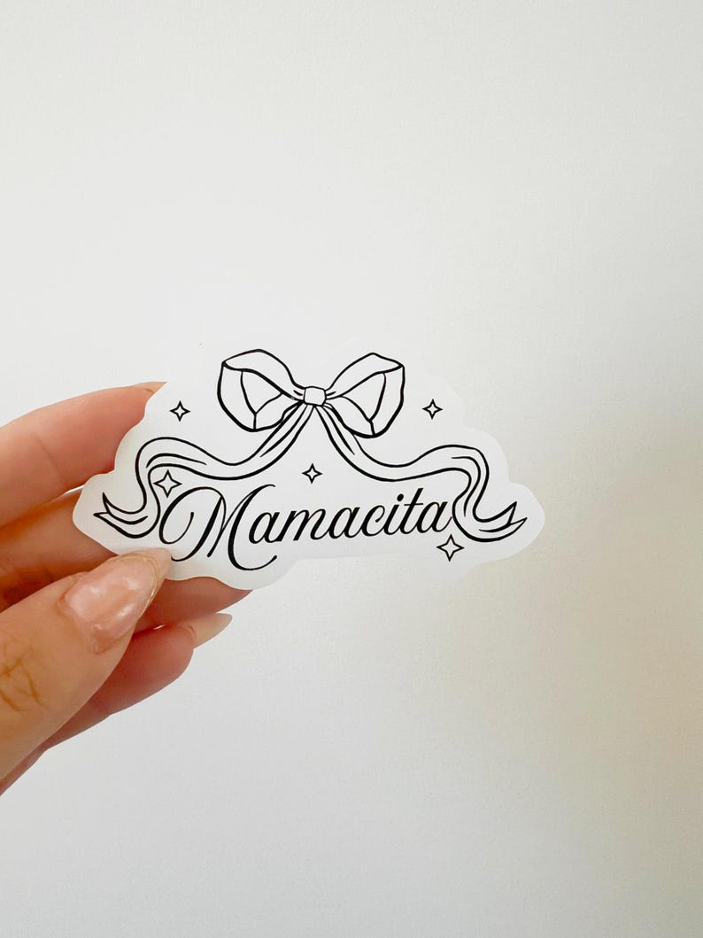 May include: A white sticker featuring a black bow and the word "Mamacita" in cursive. The design includes a ribbon and four star shapes. A decorative sticker.