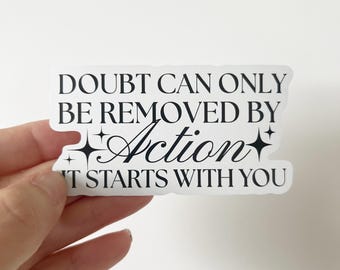 Op de afbeelding: Een witte sticker met zwarte tekst die luidt: "DOUBT CAN ONLY BE REMOVED BY Action IT STARTS WITH YOU". Het woord "Action" is in een cursief lettertype met sterretjesaccenten.