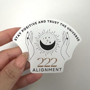 May include: A white sticker with the text "STAY POSITIVE AND TRUST THE UNIVERSE" in black, arched across the top. Below, a black crescent moon and stars are centered between two hands. The number "222 ALIGNMENT" is printed in gold.