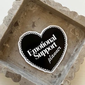 Op de afbeelding: Een zwarte sticker in de vorm van een hart met witte tekst die "Emotional Support Planner" zegt op een grijs betonnen dienblad.