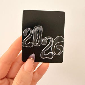 May include: A black card with the year "2020" written in a clear, bubbly font. The card is held by a hand with a gold ring on the index finger and a purple and clear nail design.