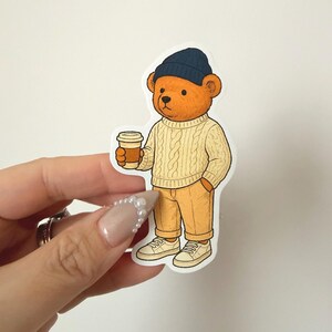 Pu&ograve; includere: Un adesivo con un orsetto dei cartoni animati con un berretto blu navy, un maglione a trecce color crema e pantaloni color cachi. L'orso tiene in mano una tazza di caff&egrave; e indossa scarpe da ginnastica bianche. L'adesivo ha un bordo bianco.