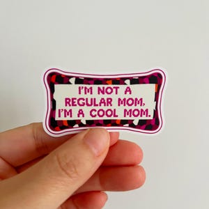 May include: A rectangular sticker with a white background and a decorative border in shades of pink, black, and orange. The sticker reads, "I'M NOT A REGULAR MOM. I'M A COOL MOM." The sticker is held by a hand.