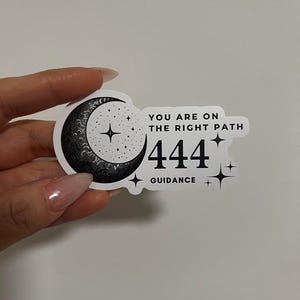 May include: A white sticker with a black crescent moon design, stars, and the text "YOU ARE ON THE RIGHT PATH 444 GUIDANCE". The sticker is held by a hand with manicured nails.