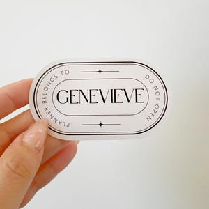 May include: An oval sticker with the name "GENEVIEVE" in a serif font, framed by a black border. The text "PLANNER BELONGS TO" and "DO NOT OPEN" are in a circular pattern around the name. The sticker is held by a hand.