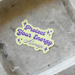Puede incluir: Una pegatina amarilla con el texto "Protect Your Energy Always" en púrpura y acentos de estrellas. La pegatina está sobre una superficie texturizada de color gris claro.