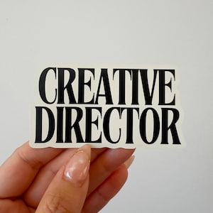 May include: A white sticker with the words "CREATIVE DIRECTOR" in bold, black capital letters. The sticker is held by a hand against a white background.